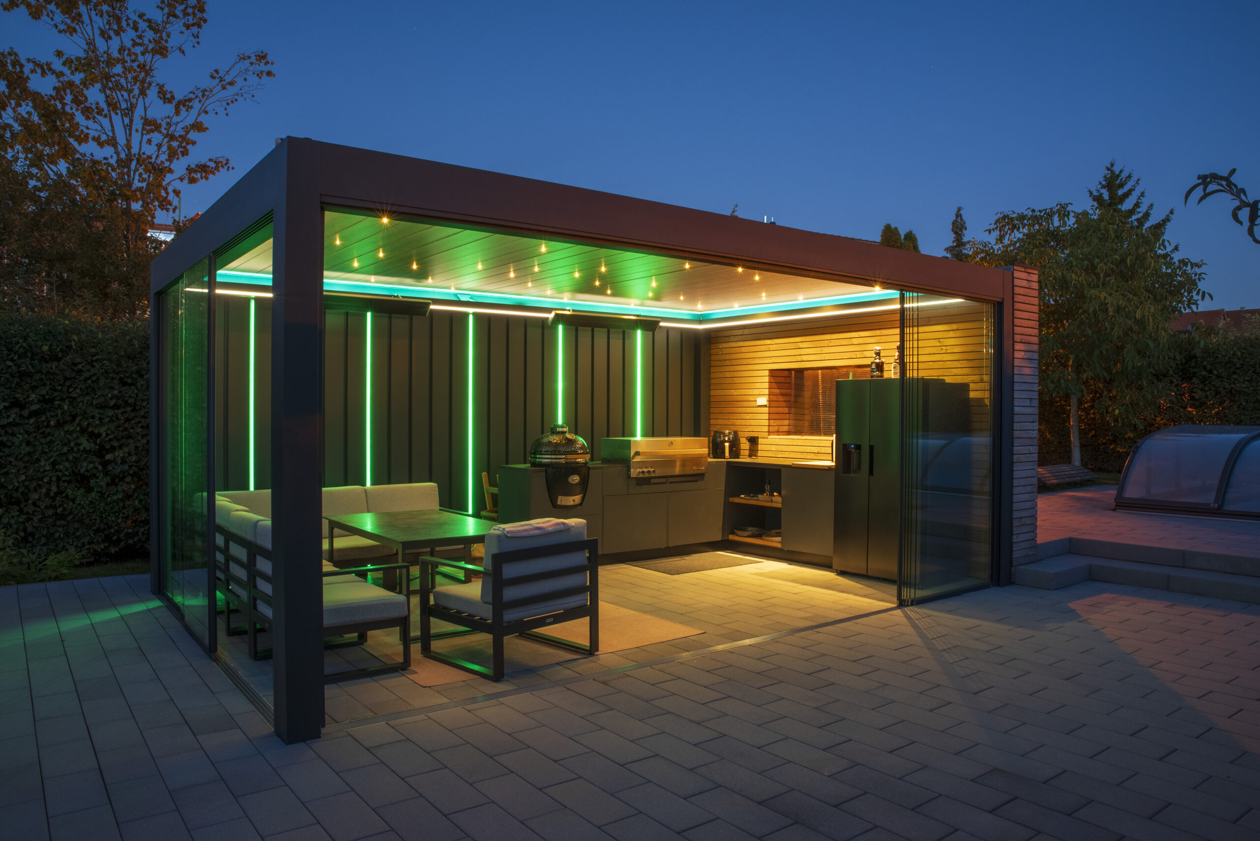 Mit Lamellendach (geschlossen) überdachte Outdoor-Küche mit großem Outdoor-Kühlschrank, Outdoor-Spüle, 2 Grills und Essecke. Grüne und gelbe LEDs sorgen am Abend für stimmungsvolle Beleuchtung.