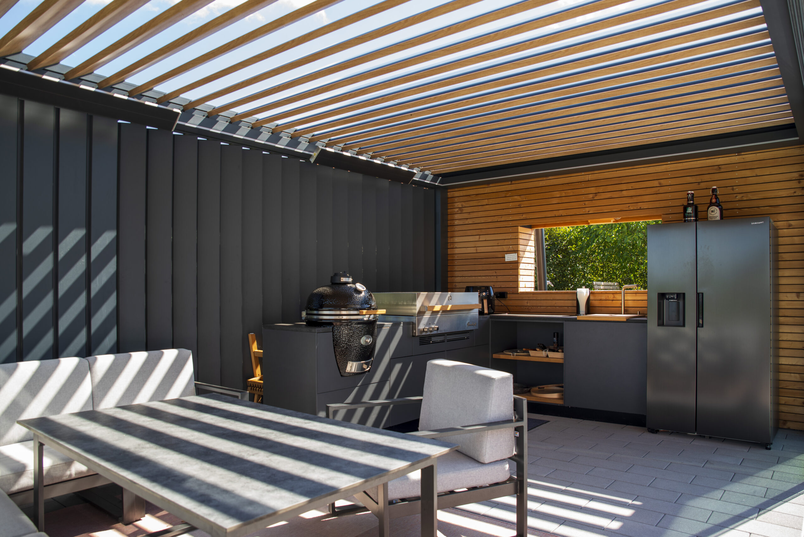 2 Grills, Outdoor-Spühle und Outdoor-Kühlschrank in einer mit Lamellen-Pergola überdachten Outdoor-Küche.