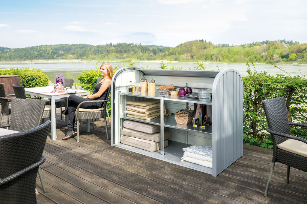 Biohort StoreMax: Die Gartenbox steht mit geöffnetem Rollladen auf der Terrasse eines Restaurants und enthält Polster, Decken und Accessoires.