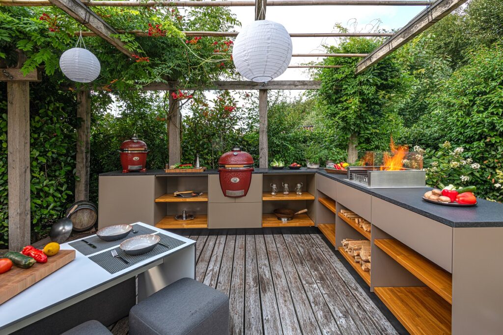 Beige Outdoor-Küche mit beigem Korpus, schwarzer Arbeitsplatte und Eichenholz-Einlegeböden sowie 2 Monolith-Kamados und einer Feuerstelle. Darüber eine Pergola.