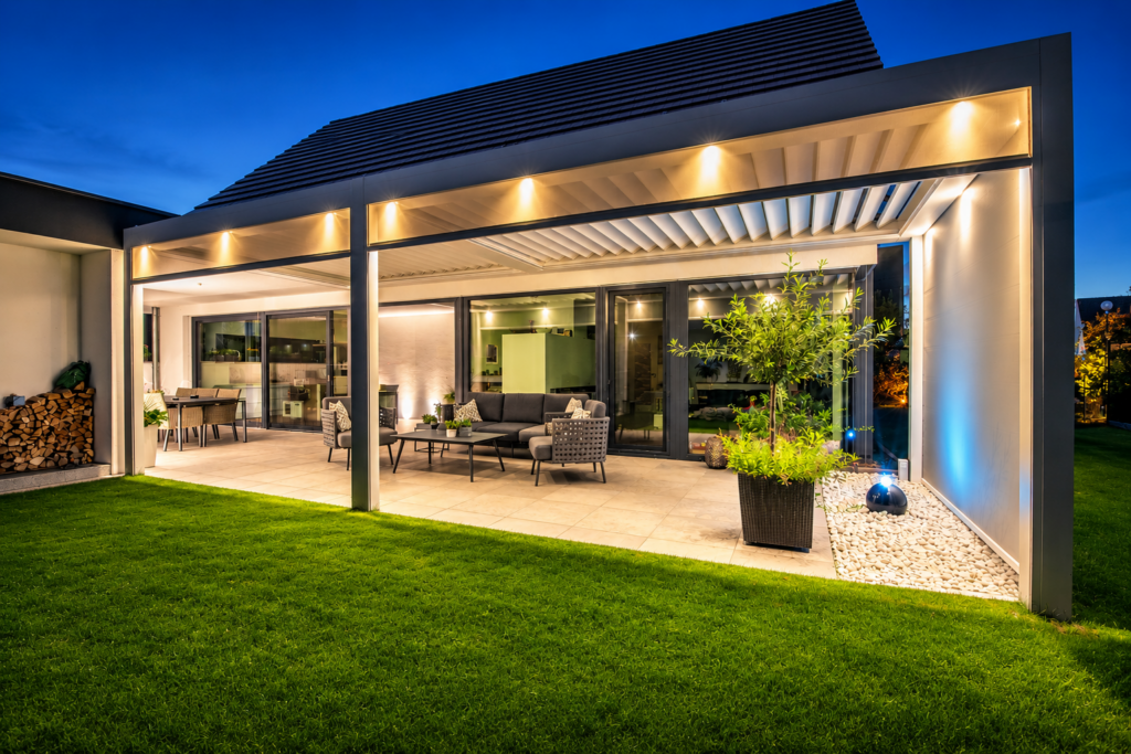 Moderne Lamellenpergola mit integrierter LED-Beleuchtung auf einer stilvollen Terrasse bei Abenddämmerung. Die hochwertige Outdoor-Lounge und der Essbereich schaffen ein exklusives Ambiente und verbinden elegantes Design mit maximalem Wohnkomfort im Freien.